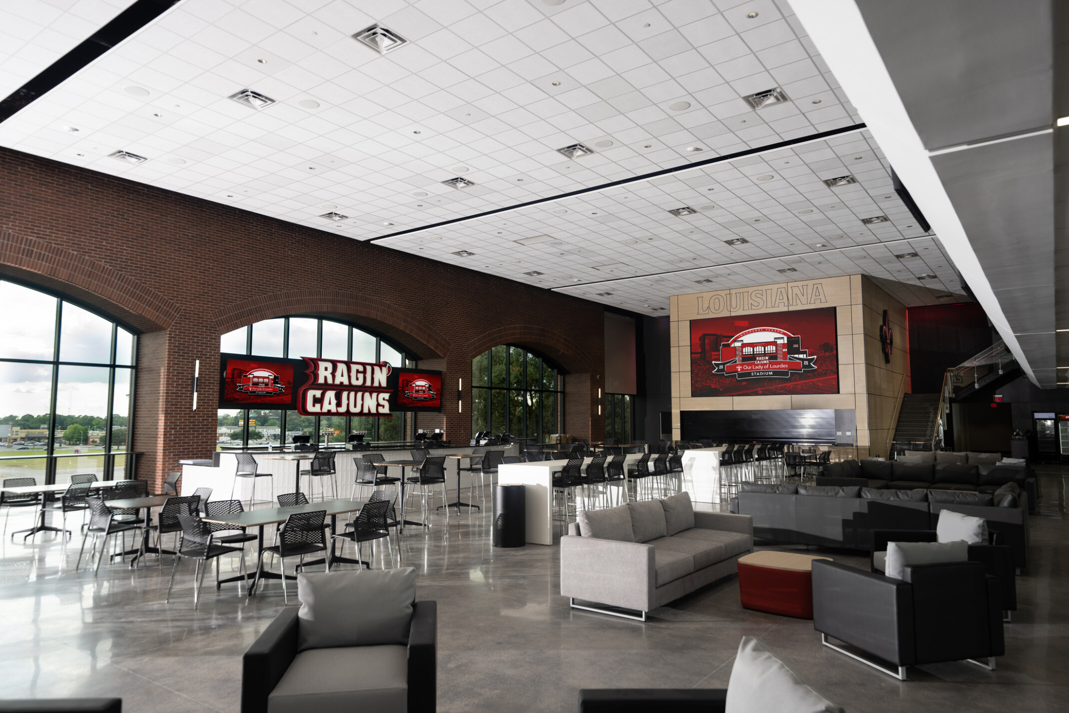 University of Louisiana Our Lady of Lourdes Stadium west bowl transformation; athletic lounging space with varied seating and bar space; red white and gray color palette with "Ragin Cajuns" sign on top of the bar