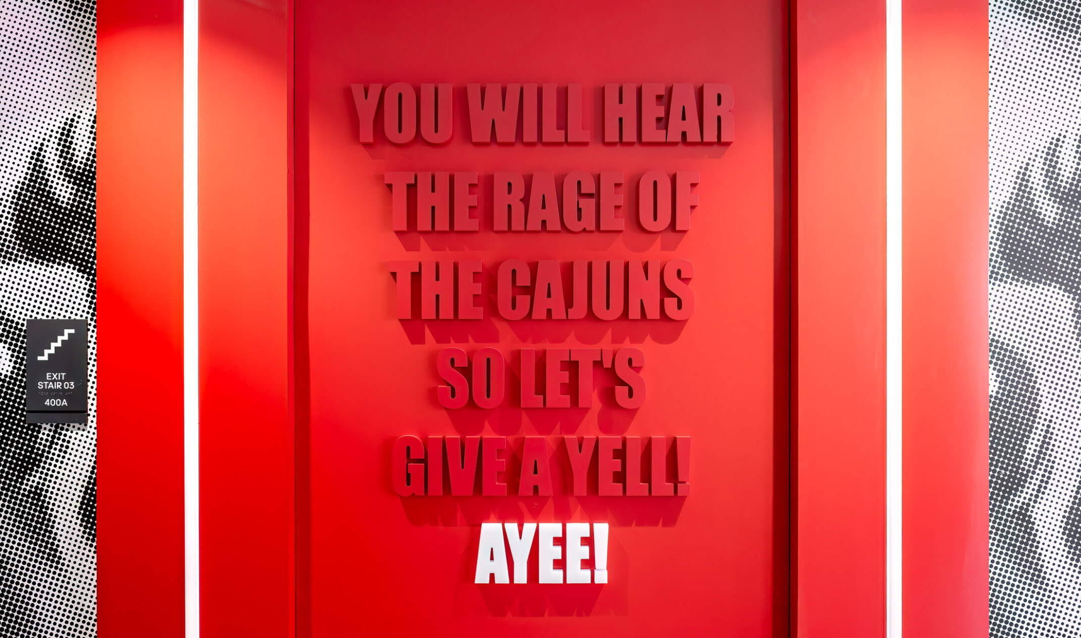 Bright red interior wall displaying the text “YOU WILL HEAR THE RAGE OF THE CAJUNS SO LET’S GIVE A YELL! AYEE!” in raised lettering, framed by illuminated vertical light strips and patterned black-and-white side panels.
