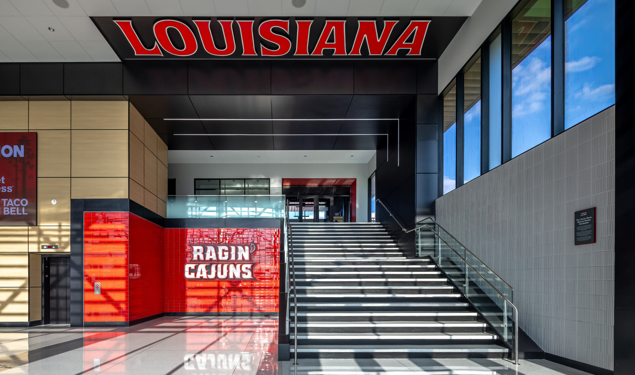 Interior stadium entrance featuring a grand staircase, illuminated “Louisiana” signage overhead, Ragin’ Cajuns branding, glass railings, and daylight streaming through floor-to-ceiling windows.