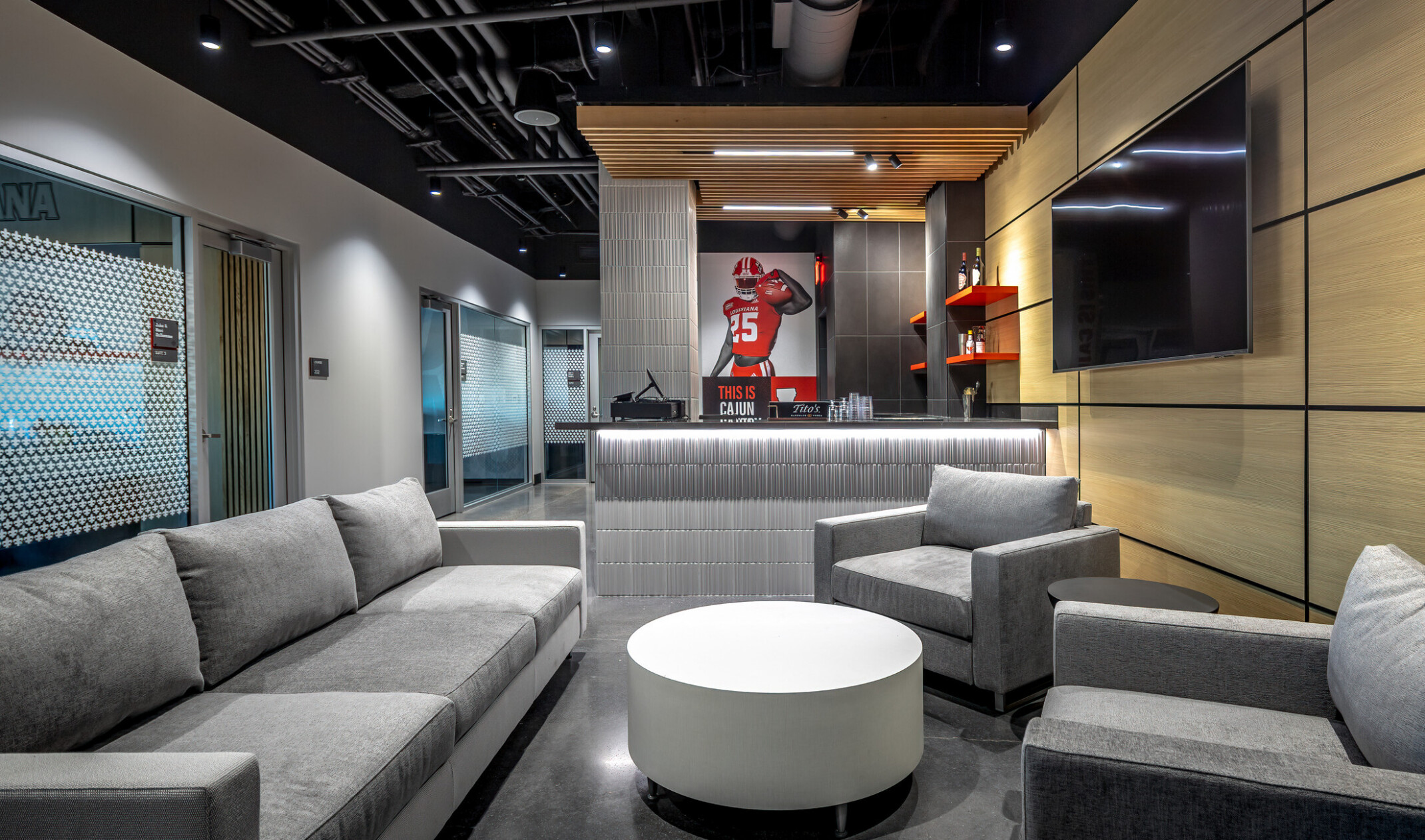 Modern premium suite lounge featuring gray seating, a round coffee table, a private bar area, wood-paneled walls, and a large television. A Ragin’ Cajuns football graphic is displayed at the back of the room.