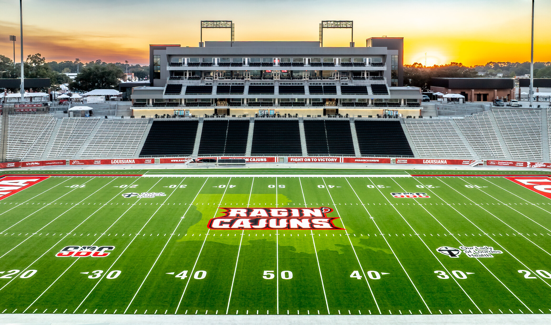 Wide view of an empty football stadium at sunset, featuring a green field with the “Ragin’ Cajuns” logo at midfield and grandstands in the background.