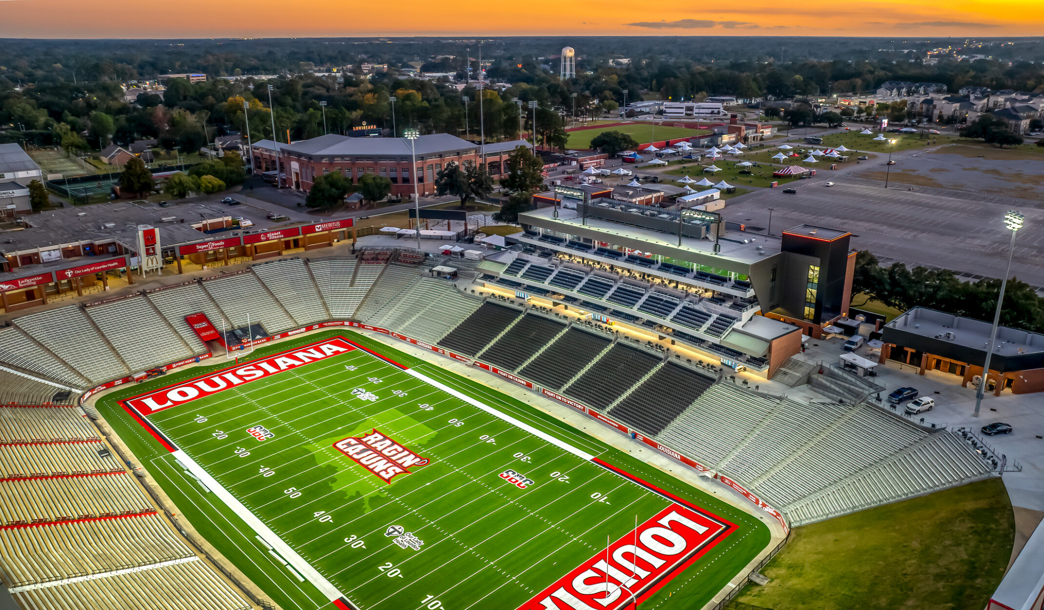 Centered field-level view of a football stadium during the day, showing the “Ragin’ Cajuns” logo at midfield, yard line markings, and a multi-level press box behind rows of empty seats.