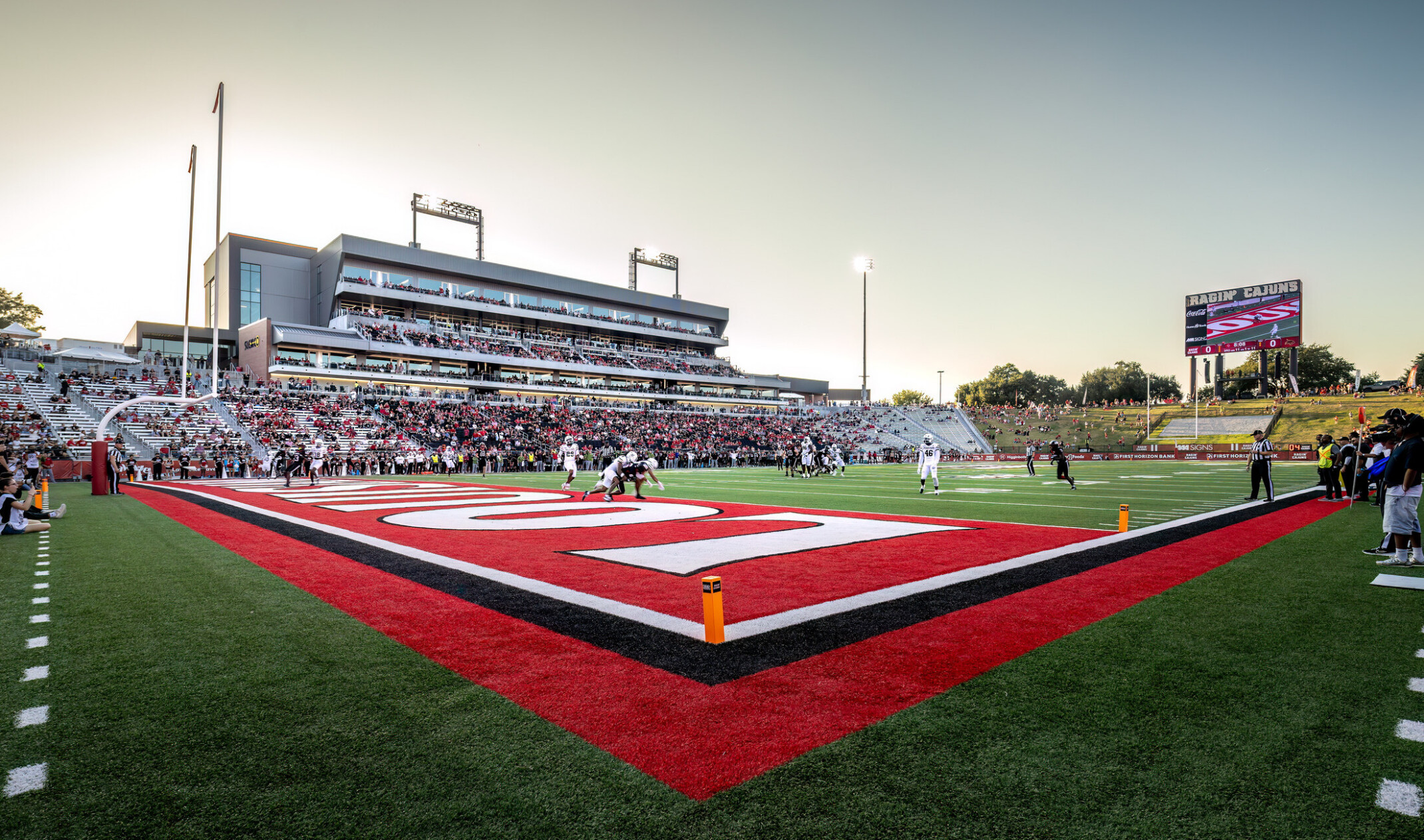 Football game in progress at a large outdoor stadium, viewed from the corner of a red end zone with players on the field and spectators filling the stands under an evening sky.