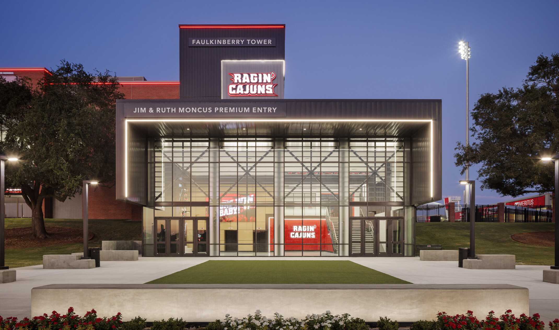 Evening view of the Faulkinberry Tower premium entrance with illuminated Ragin’ Cajuns branding, a glass façade, landscaped plaza, and stadium lighting in the background.