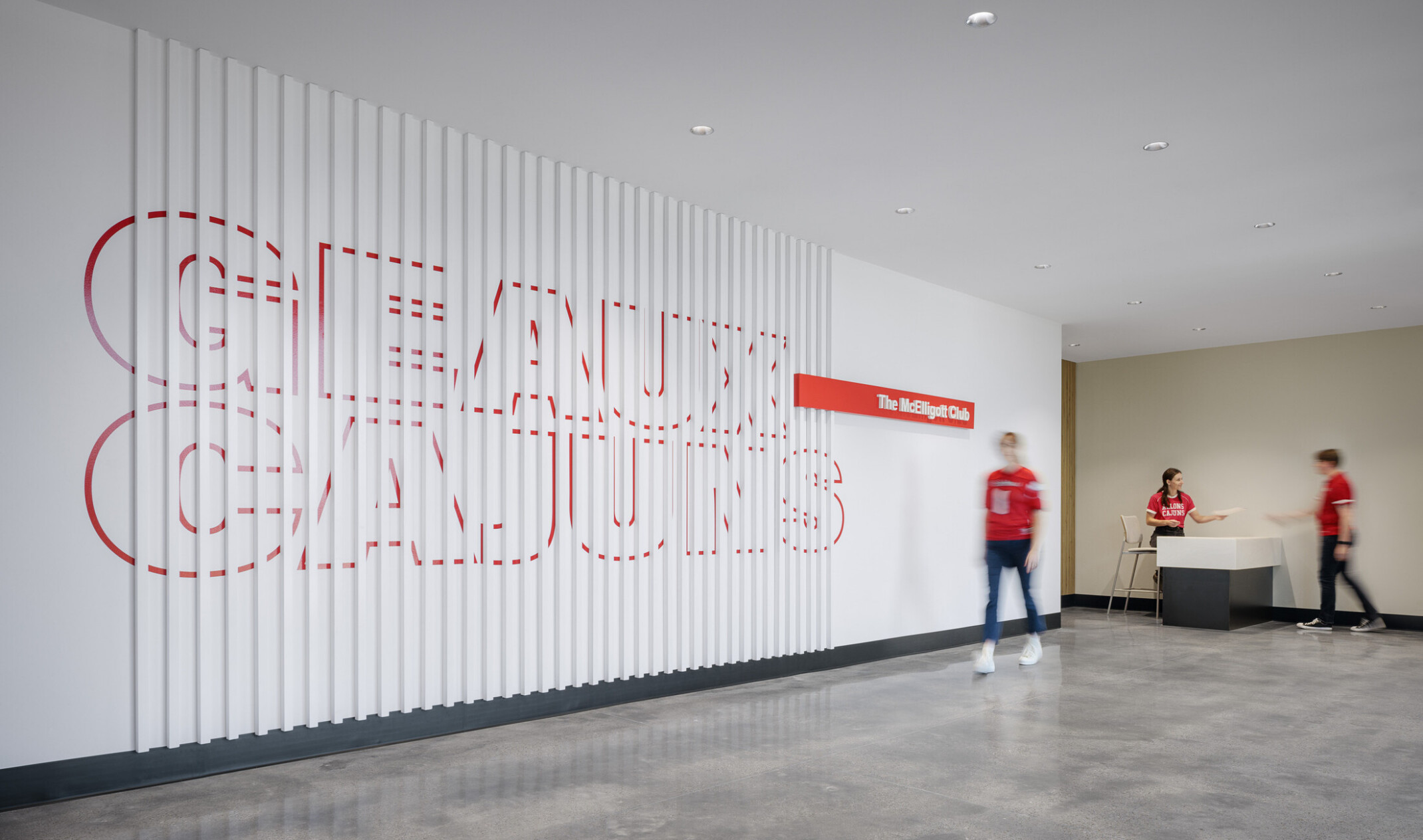Modern stadium club entrance featuring a large decorative wall with the words “Ragin’ Cajuns” integrated into vertical white panels. A reception desk labeled “The McElligott Club” sits at the end of the corridor, with visitors walking through the space