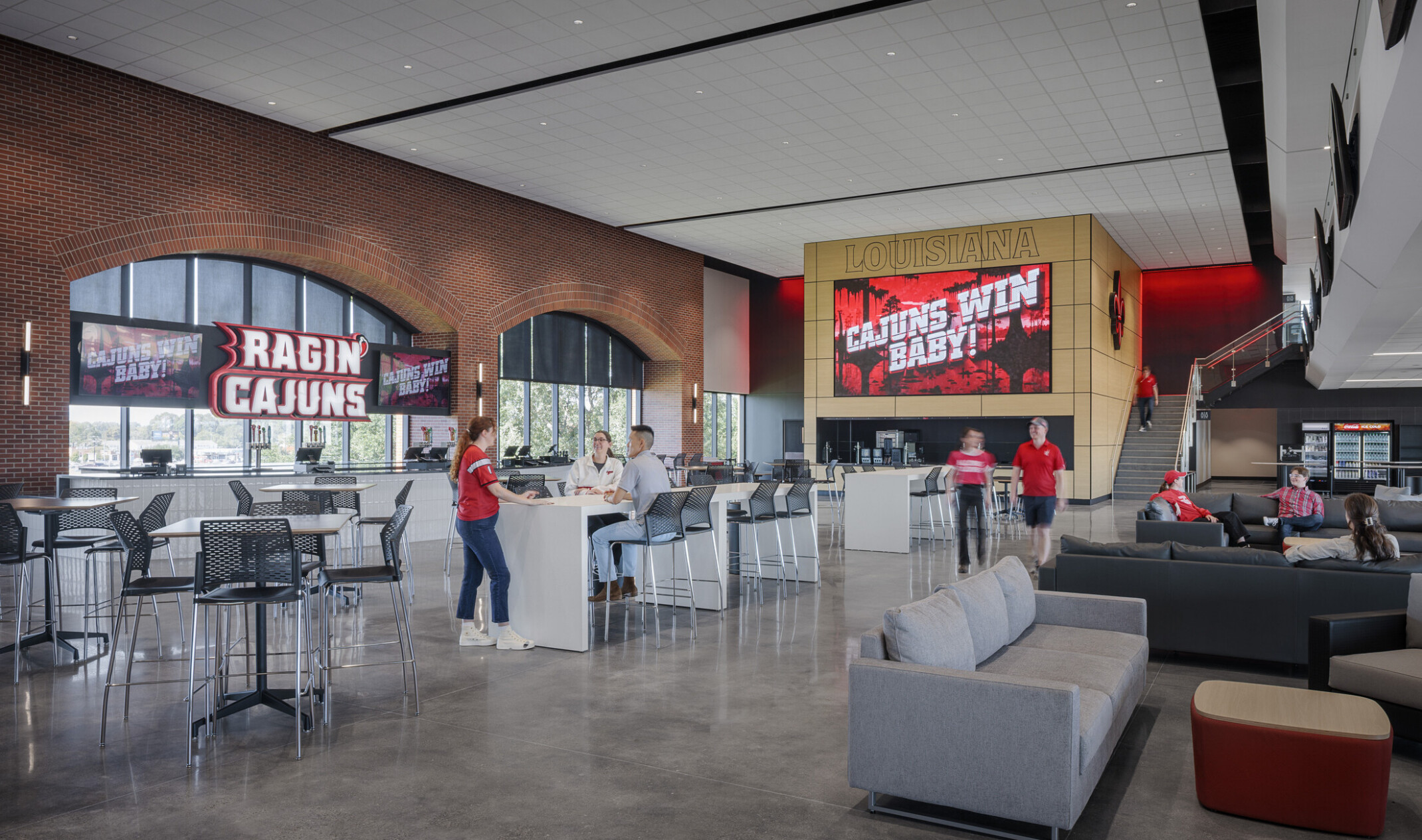 Spacious stadium interior lounge with modern seating, tables, and large digital screens displaying “Ragin’ Cajuns,” set against brick walls and tall arched windows