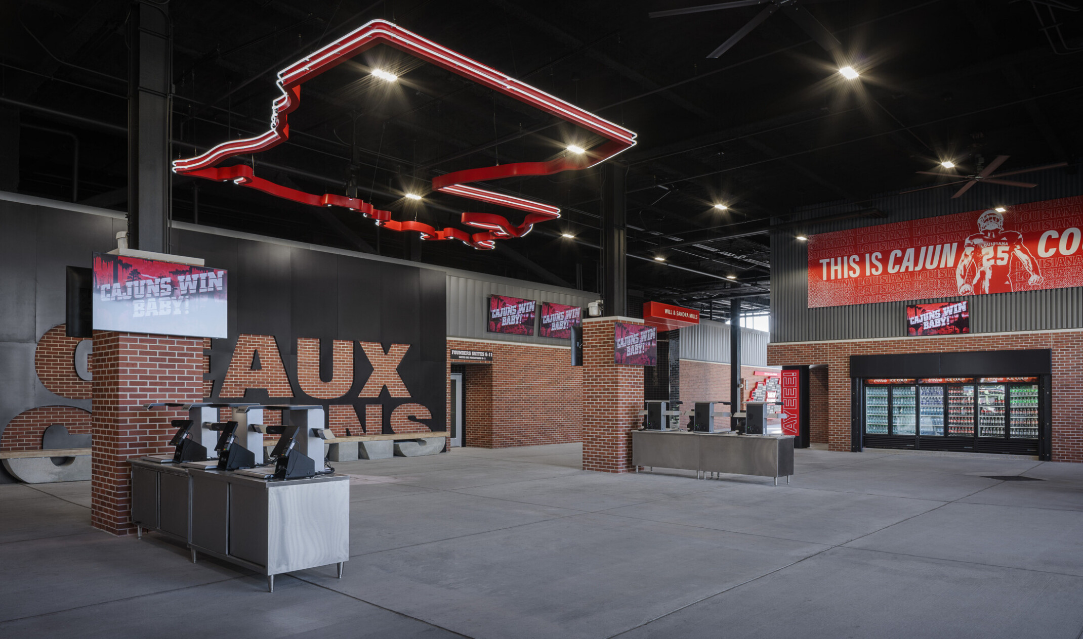 Large club-level gathering space inside a football stadium with lounge seating, dining tables, concession areas, and prominent Ragin’ Cajuns branding. Visitors socialize throughout the open room beneath high ceilings and large digital displays