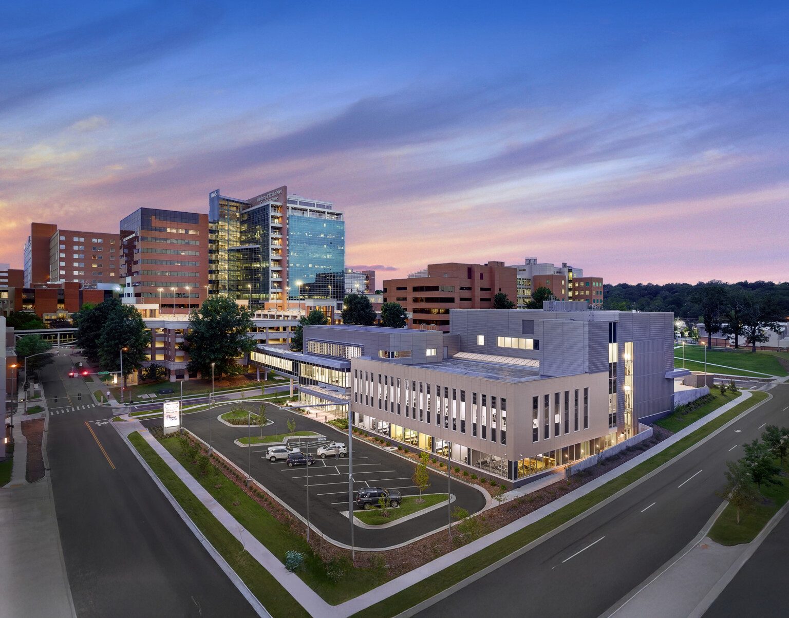 A modern hospital complex at dusk featuring multiple buildings, including a prominent glass-fronted structure and several brick buildings. The sky transitions from blue to pink, and the complex is surrounded by roads, parking areas with cars, and landscaped greenery.