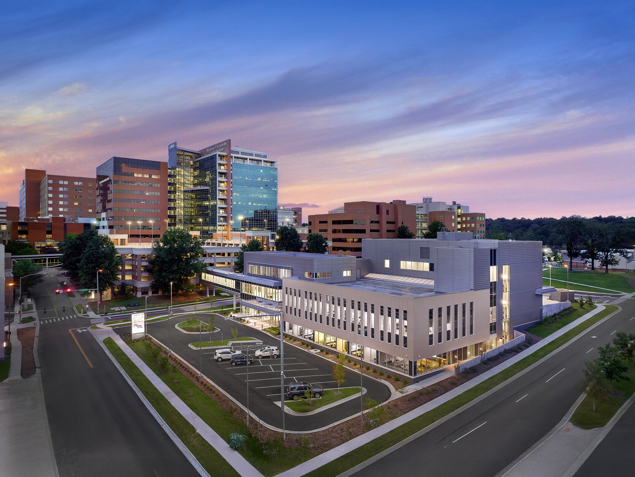 A modern hospital complex at dusk featuring multiple buildings, including a prominent glass-fronted structure and several brick buildings. The sky transitions from blue to pink, and the complex is surrounded by roads, parking areas with cars, and landscaped greenery.