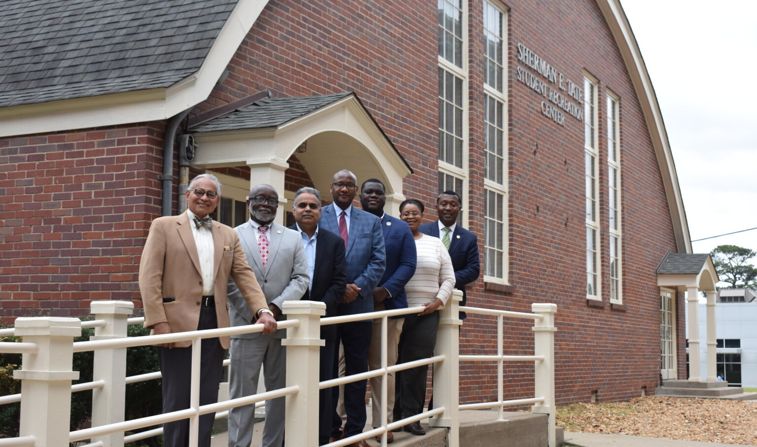 people in suits pose for photo outside a red brick building, arched roof and windows on a ramp with white metal railing, people in suits pose for photo outside a red brick building, arched roof and windows on a ramp with white metal railing,