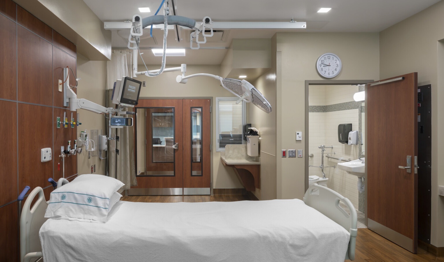 renovated hospital room with soothing dark woods, adjustable bed, IV stands, cardiac machines, creamy ivory walls and dimmable recessed lighting