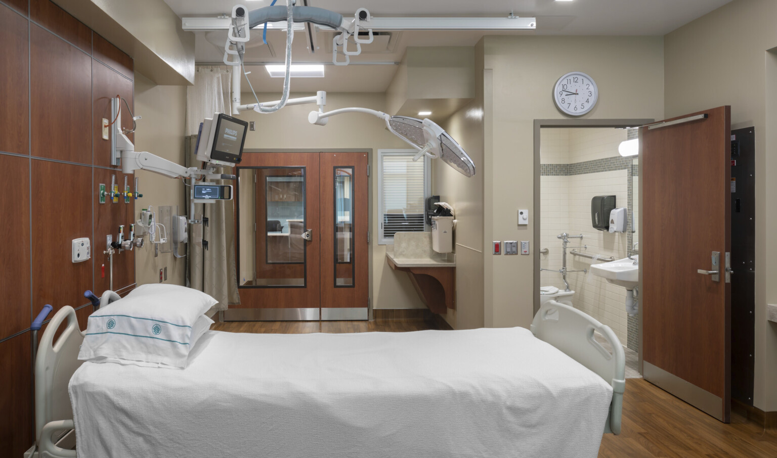 Hospital bed with equipment hanging overhead, brown wood accents on walls and doors of beige room with private bathroom Hospital bed with equipment hanging overhead, brown wood accents on walls and doors of beige room with private bathroom