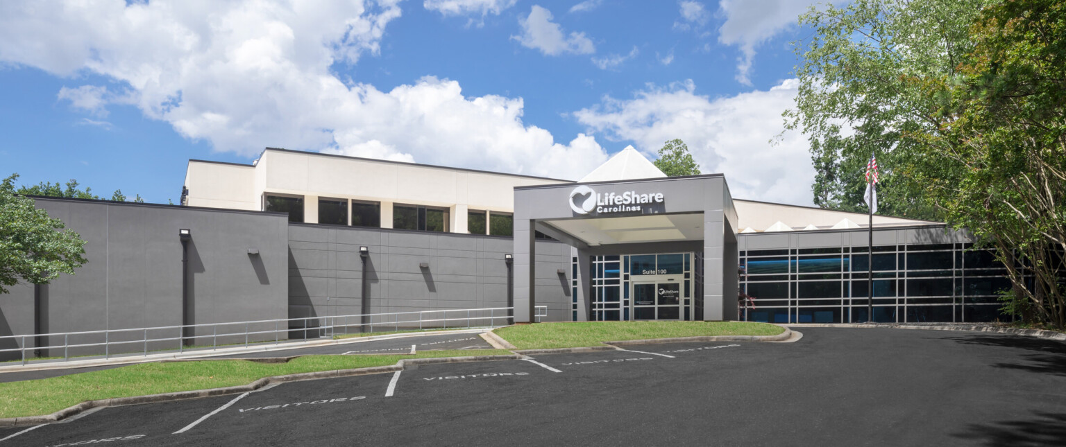 Exterior of a hospital with a black top parking lot, large trees and a white lifeshare carolinas sign above a carport