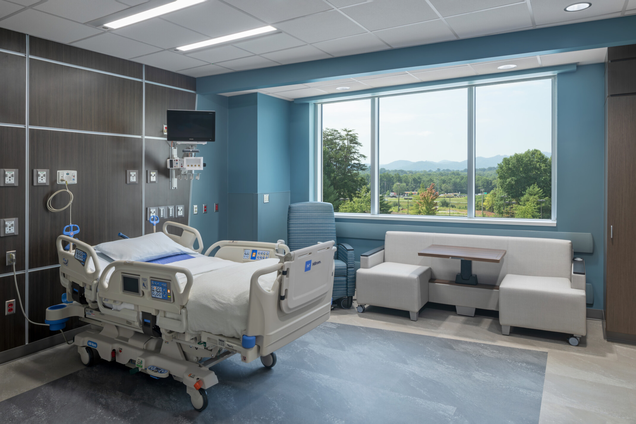 Modern hospital room with a bed, medical equipment, and a seating area by a large window