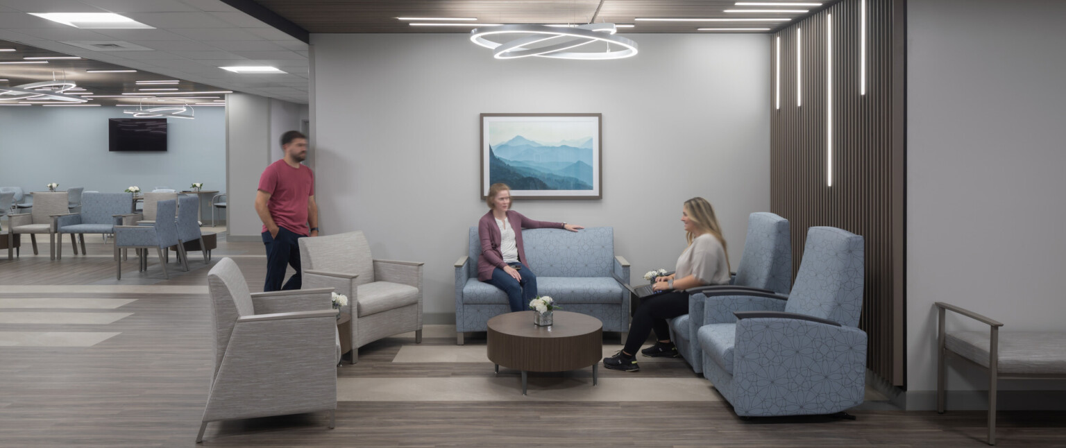 Patient waiting area filled with people sitting in blue chairs with modern circular lighting hanging from the ceiling