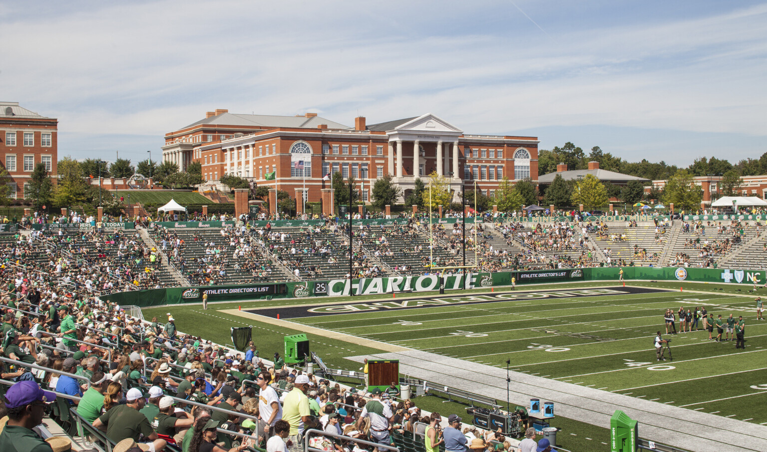 View from stands of end zone with Charlotte written across. Bleacher seats behind green wall, brick and metal fence at back
