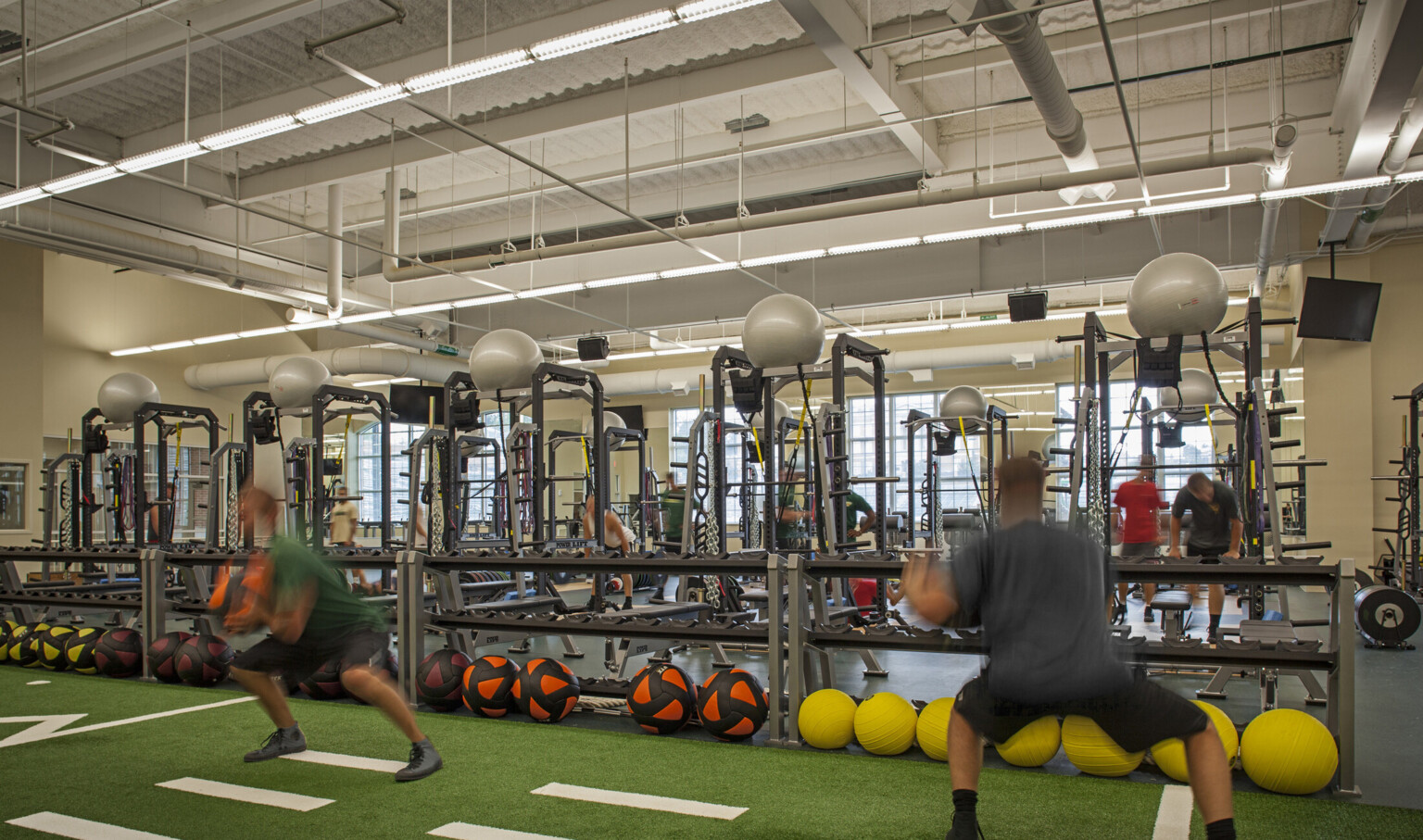 Turf floor space with yardage marked lined with half wall weight shelves in front of training equipment in cream color room