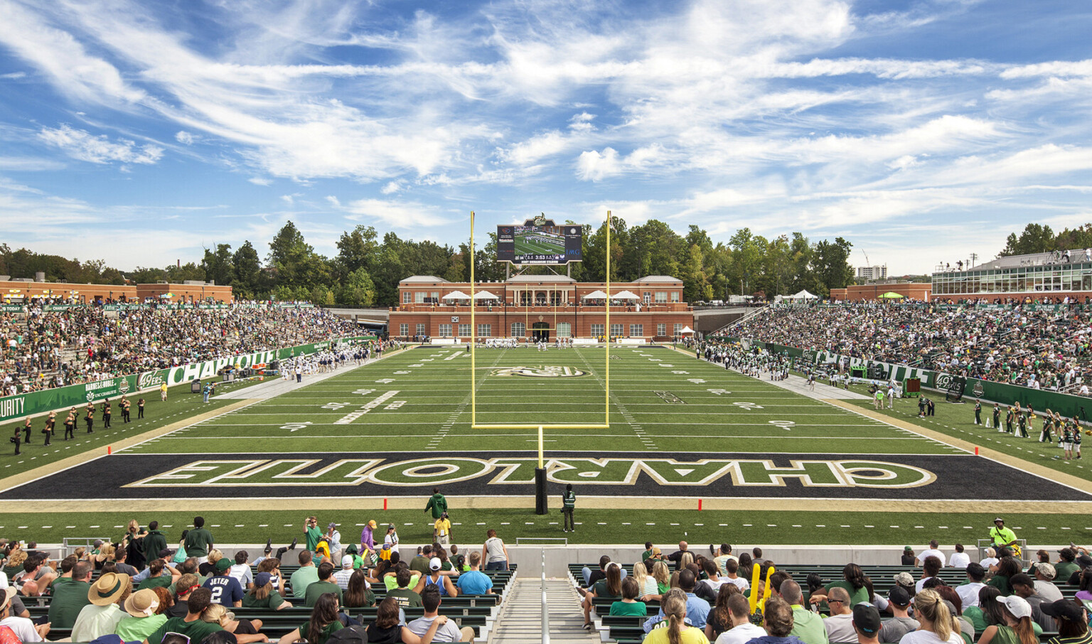 View of field from stands behind goal post looking towards brick building. Jumbotron located above 2nd floor of building