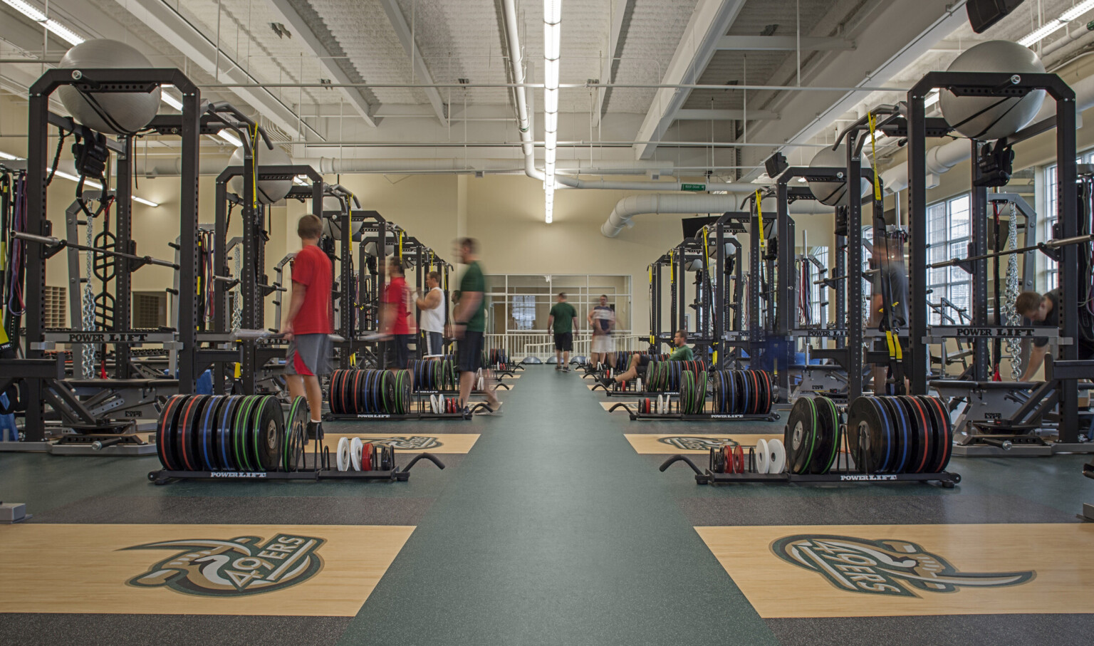 Weight room in training facility with equipment. Window lined cream colored walls under white ceiling with exposed ductwork