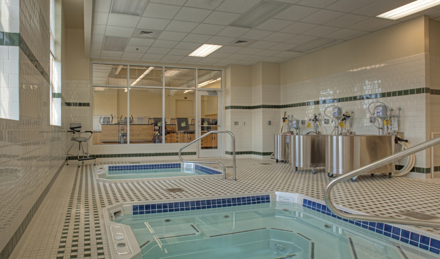 2 recovery pools in green and white tiled room with windows looking to previous room. Silver tubs along right wall