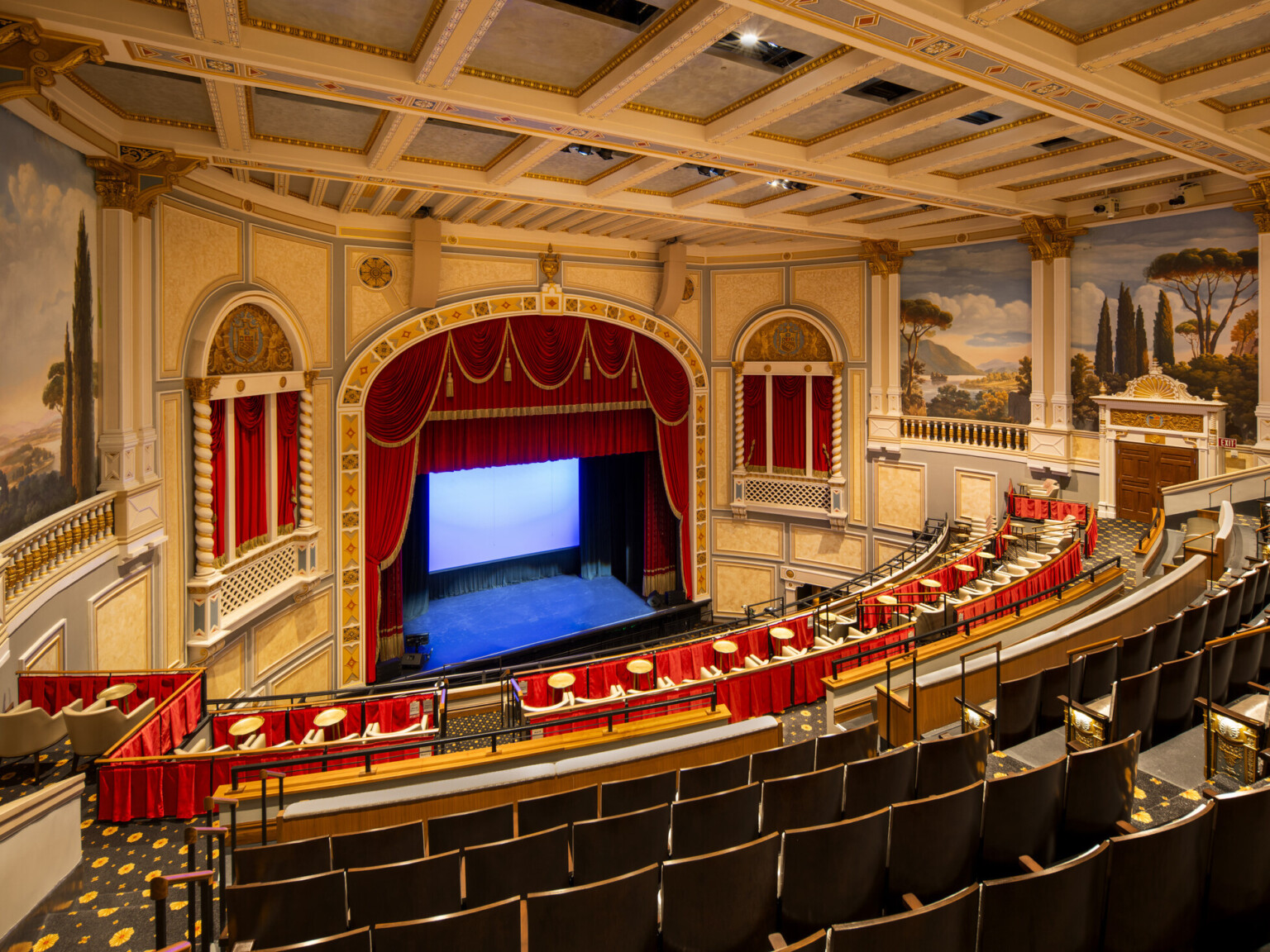 Interior of ornate theater with red velvet curtains, classical murals, and balcony seating; a private boxes with curtains