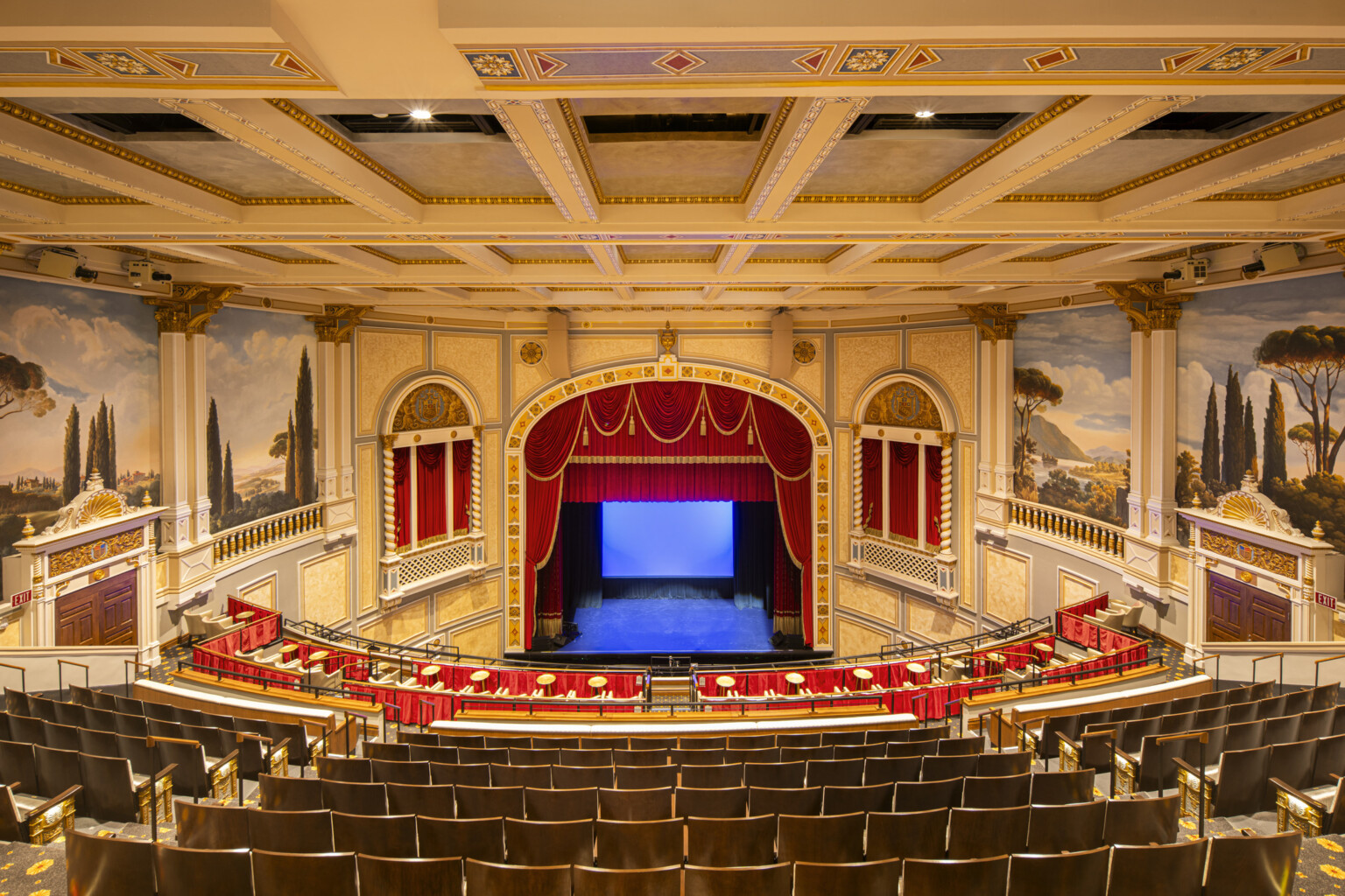 Interior of ornate theater with red velvet curtains, classical murals, and balcony seating; a private boxes with curtains