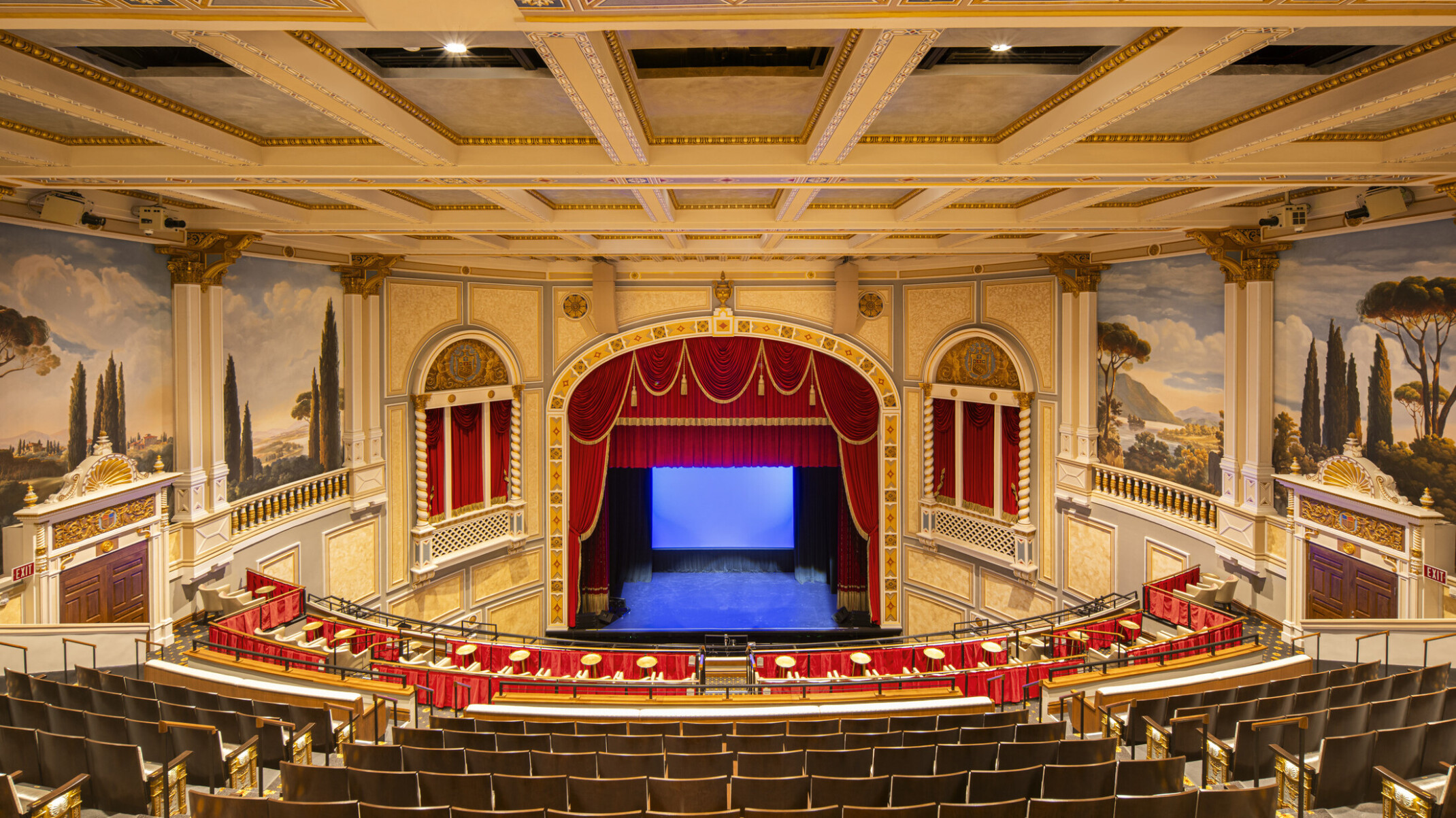 Interior of ornate theater with red velvet curtains, classical murals, and balcony seating; a private boxes with curtains