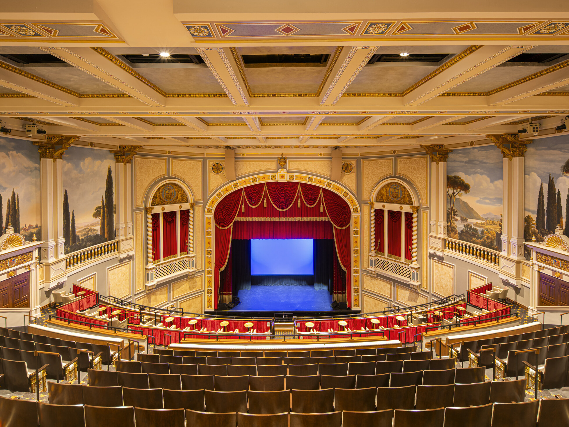 Interior of ornate theater with red velvet curtains, classical murals, and balcony seating; a private boxes with curtains