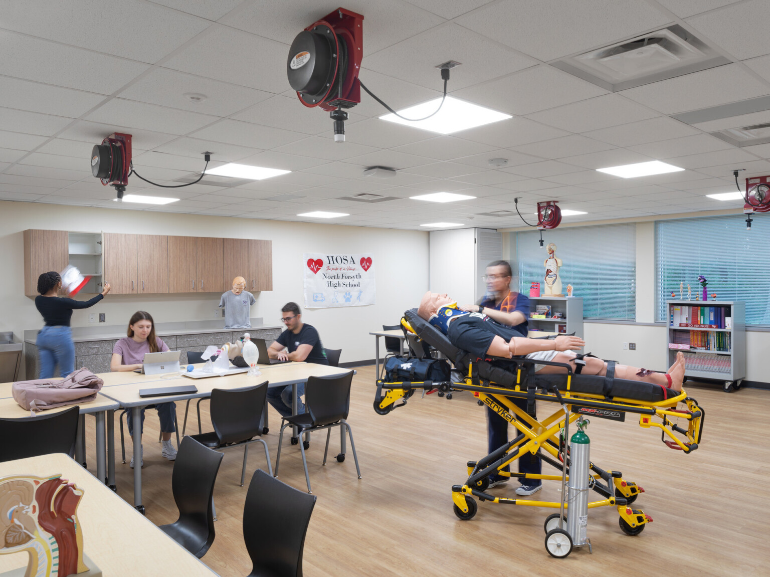 EMT classroom with natural wood floors filled with students at tables surrounding a student working on a patient on a gurney
