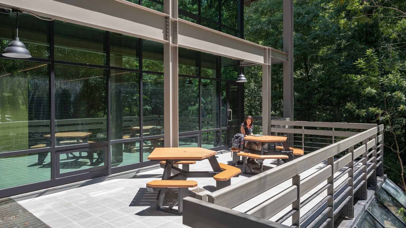 Glass building windows with brown beams and railings with picnic tables surrounded by nature