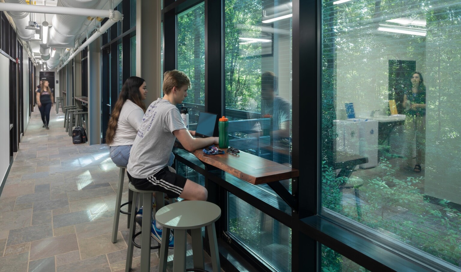 Students working on laptops in a hallway with windows, brown floors, and lots of natural lighting