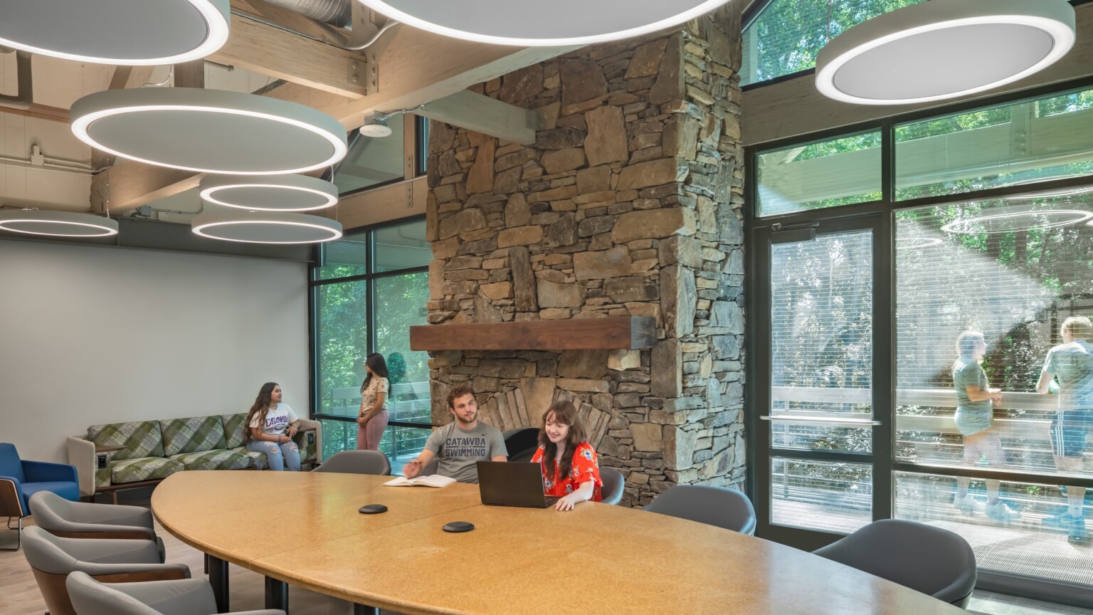 Common room, tall ceiling with wood beams accents, round pendant lights, stone fireplace in higher education setting, floor to ceiling windows