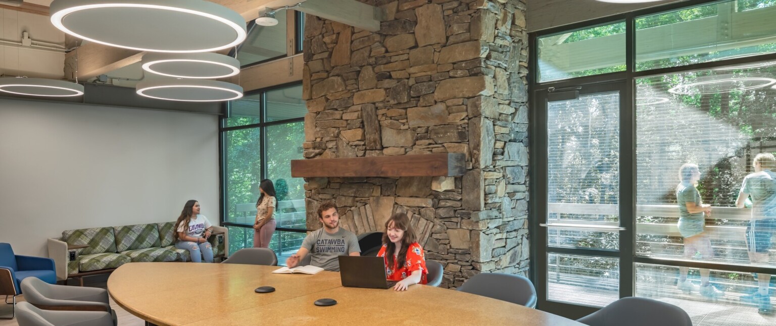 Common room, tall ceiling with wood beams accents, round pendant lights, stone fireplace in higher education setting, floor to ceiling windows