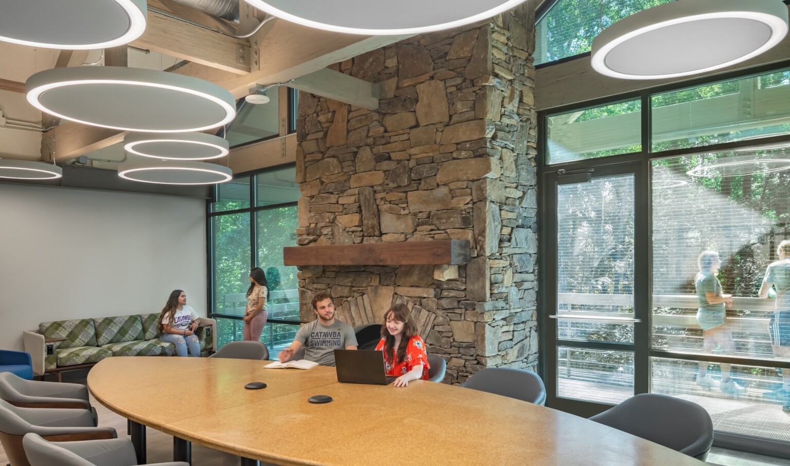 Common room, tall ceiling with wood beams accents, round pendant lights, stone fireplace in higher education setting, floor to ceiling windows