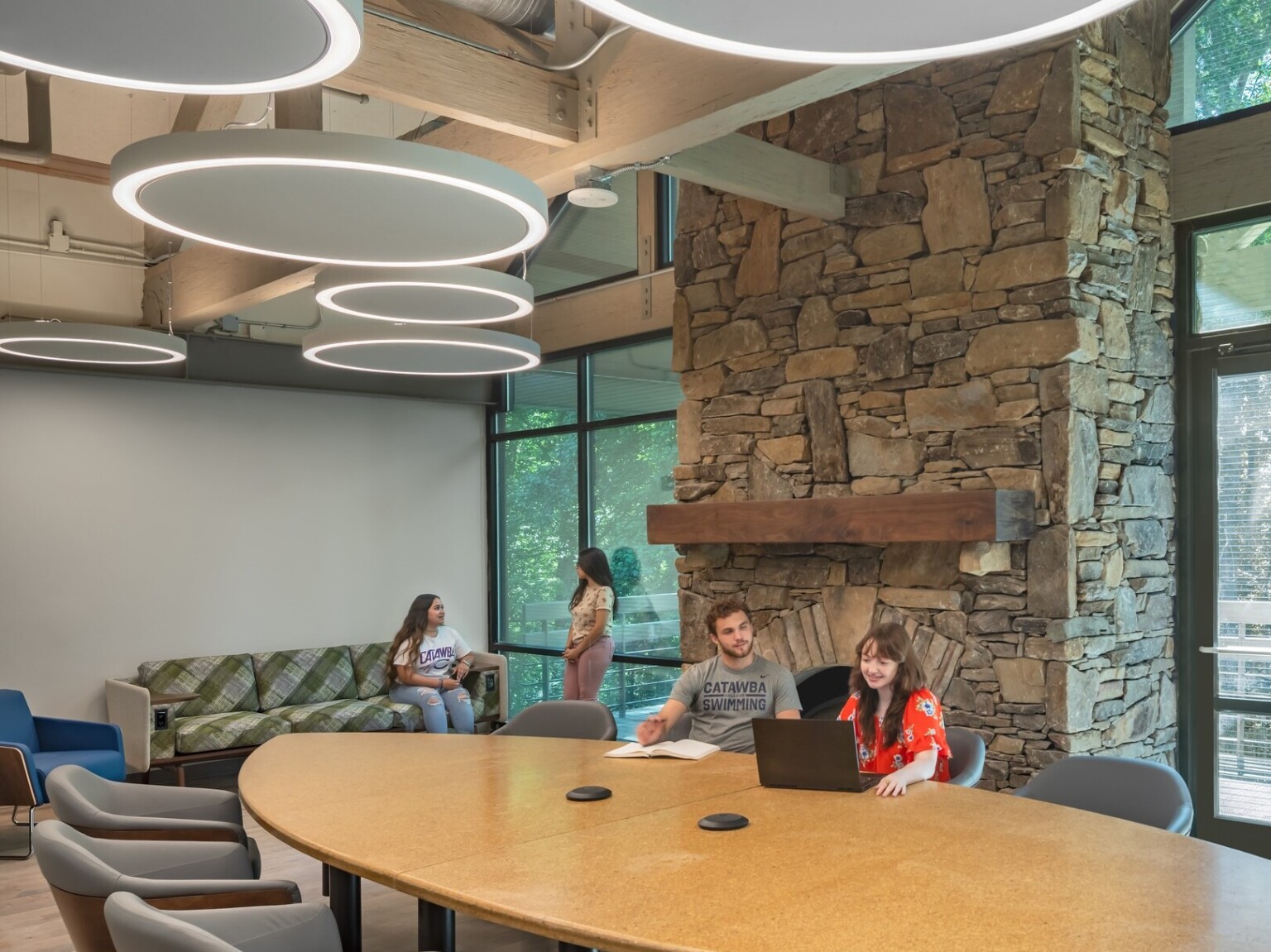 Common room, tall ceiling with wood beams accents, round pendant lights, stone fireplace in higher education setting, floor to ceiling windows