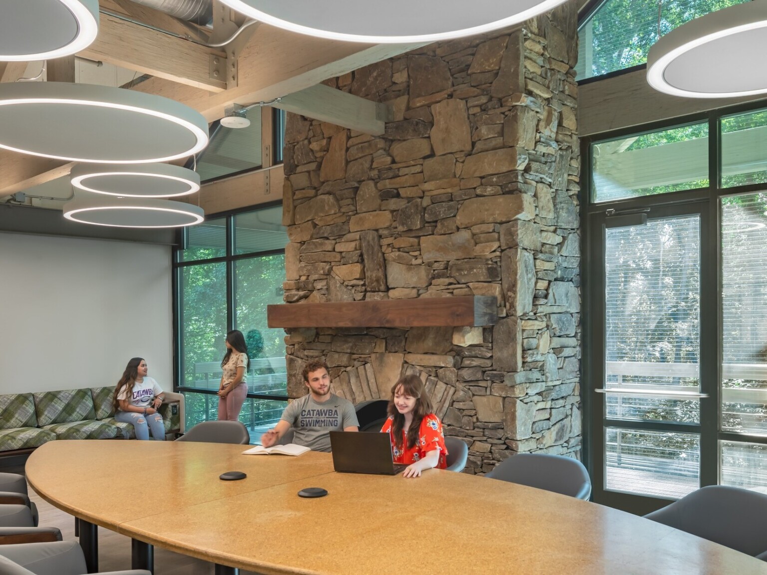 Common room, tall ceiling with wood beams accents, round pendant lights, stone fireplace in higher education setting, floor to ceiling windows