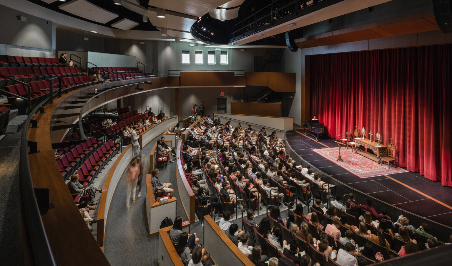 Double height auditorium theater with rounded rows of seating facing stage with red curtains, brown acoustic panels above