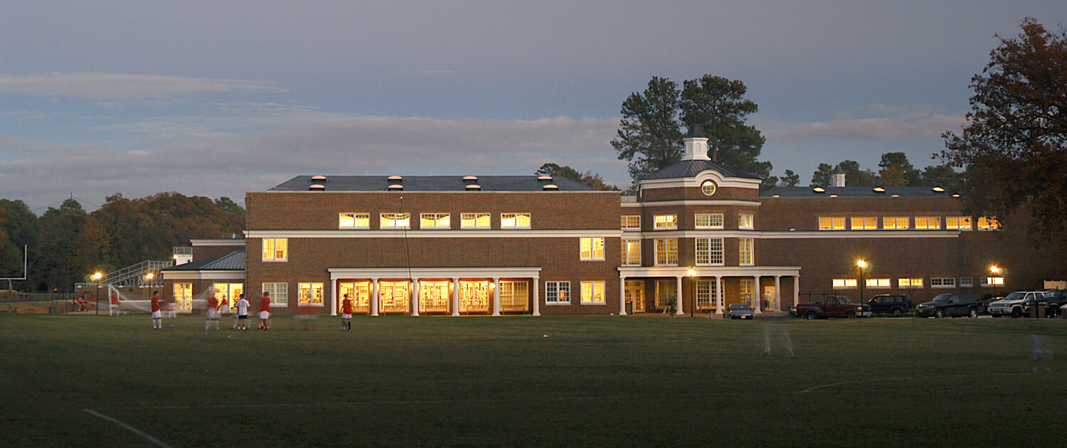 Multistory brick building behind kids playing sports with brightly lit windows with the nighttime sky in the background