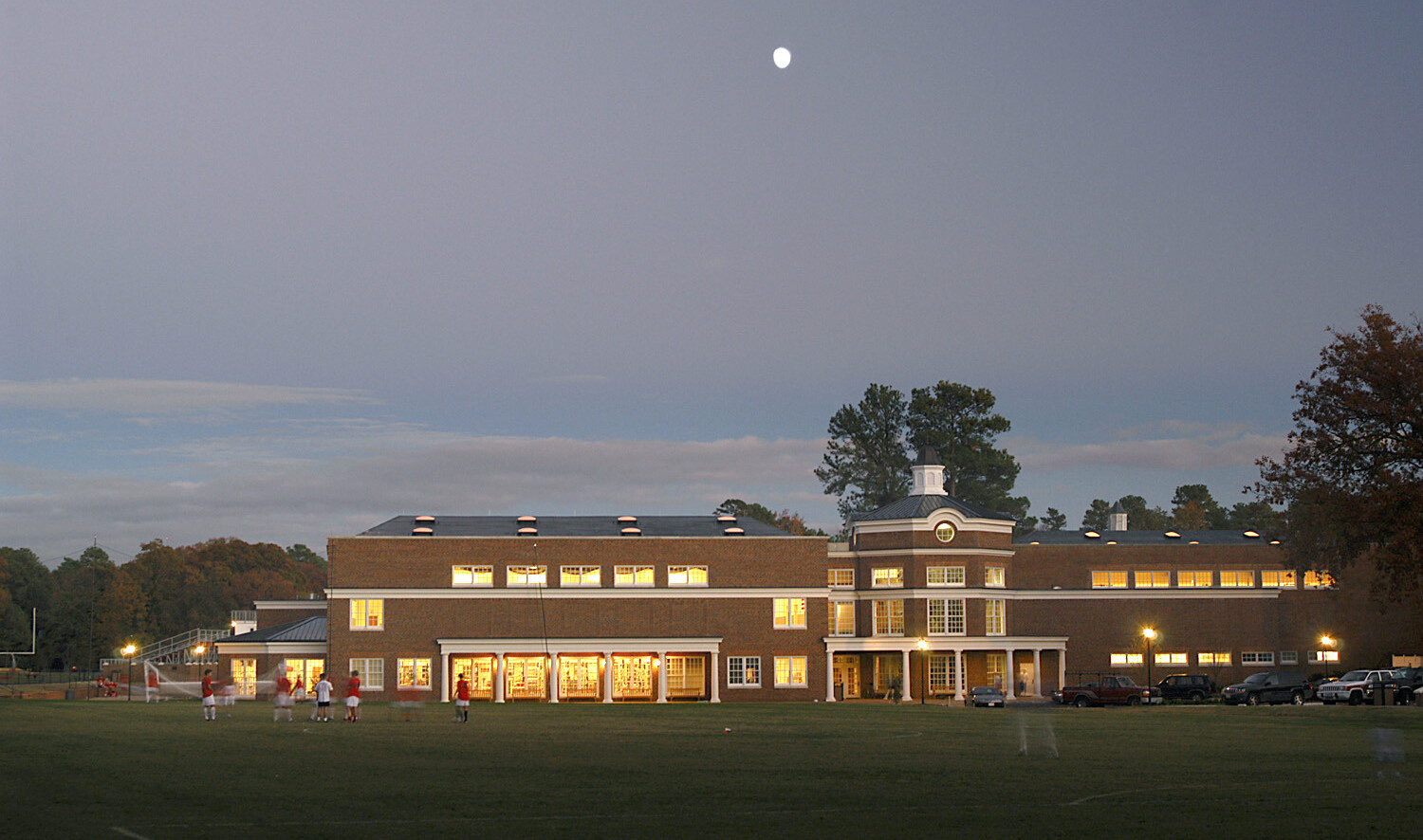 Multistory brick building behind kids playing sports with brightly lit windows with the nighttime sky in the background