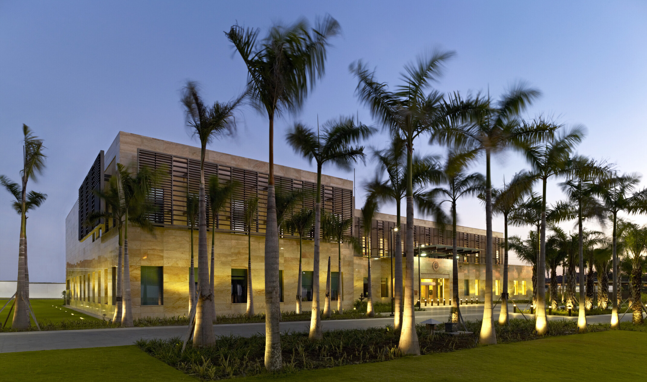 a stone building with brise soleil panels on second story windows. uplights illuminate ground floor, windows, and palm trees