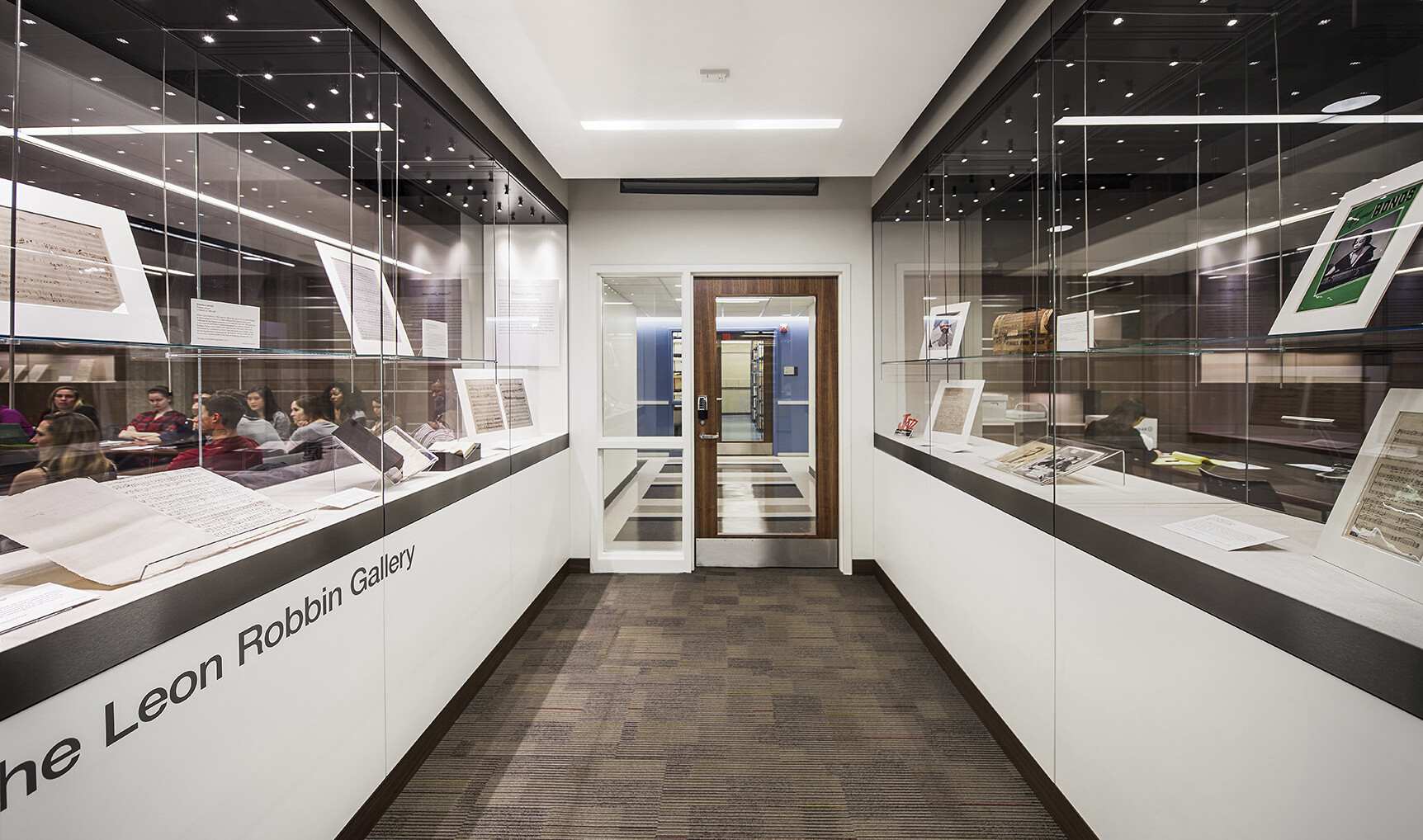 hallway of an academic library building with students seen through glass on the left and awards, plaques, and frames through glass on the right