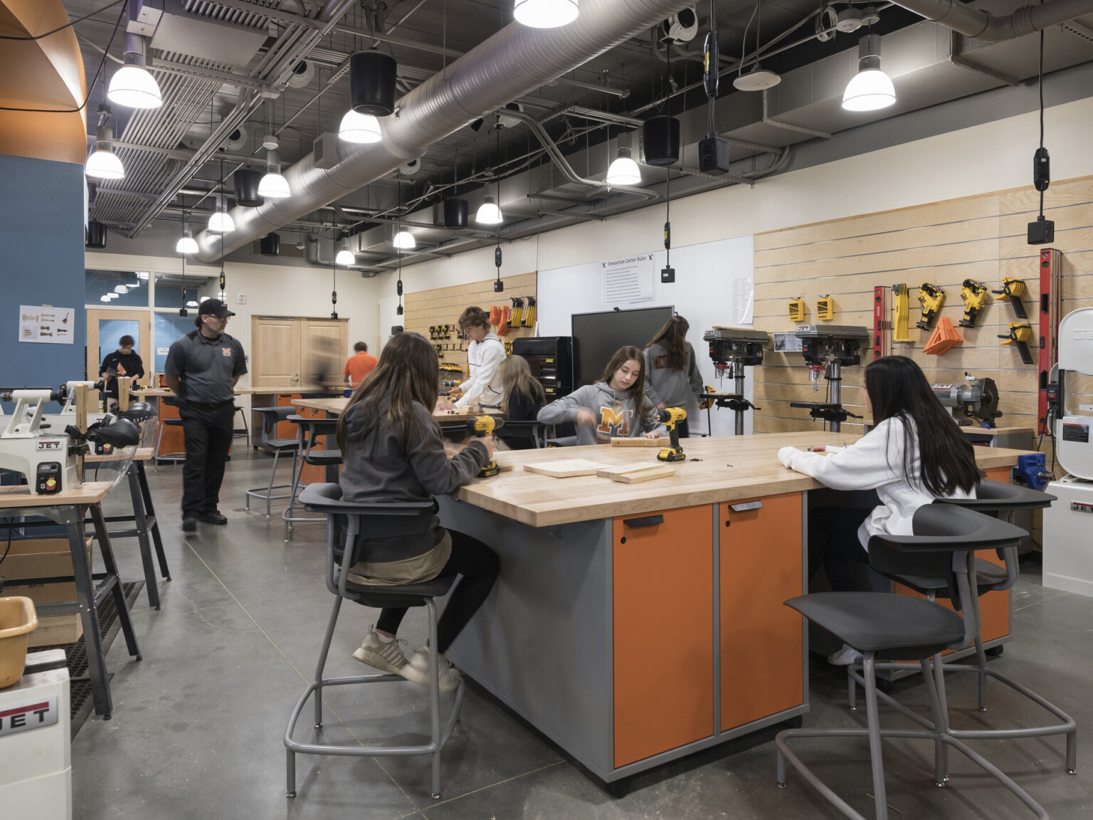 students working at orange workbenches on computers in wood paneled wood shop with tools along back wall and power cords hanging above