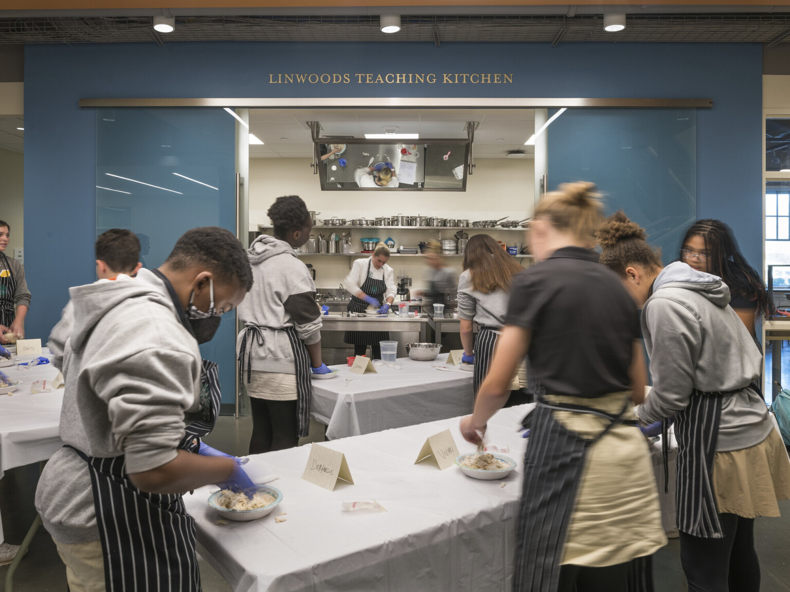 students mixing ingredients of a recipe on group work tables in the teaching kitchen, deep teal walls with overhead lighting, windows provide natural light