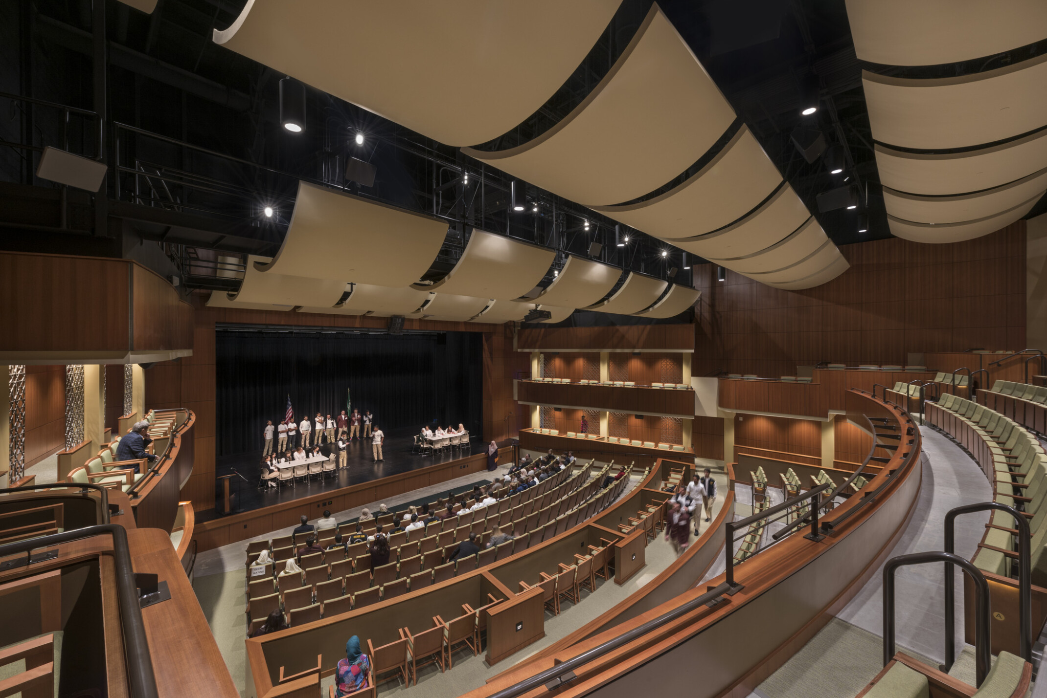 550-seat performing arts theater, view from opera boxes, suspended sound baffling, stage lighting, warm wood paneling on walls, partitions and seating, people on the stage appear to be debating or having a spelling bee