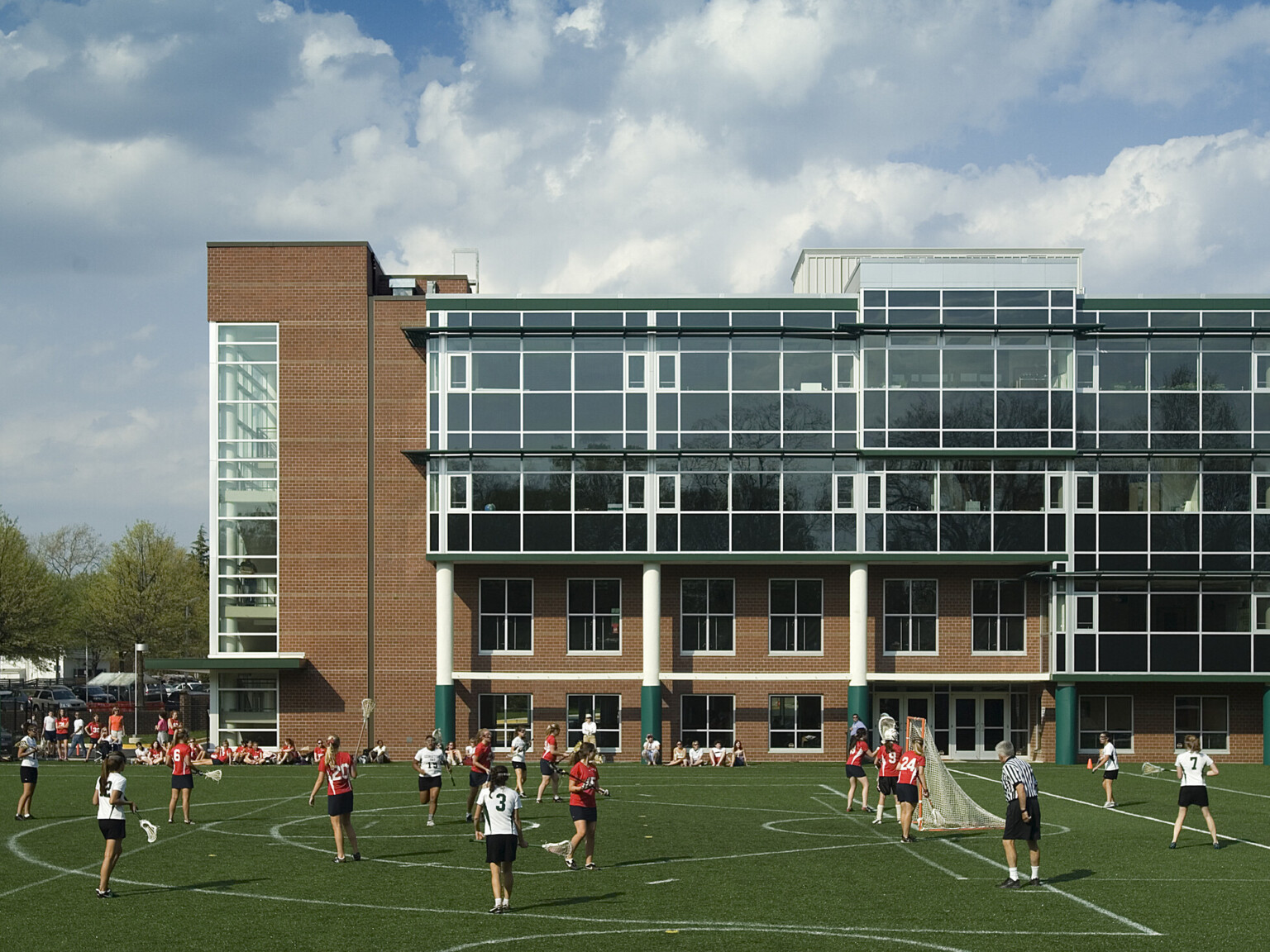 Lacrosse field filled with players in front of a multistory brick building filled with glass windows