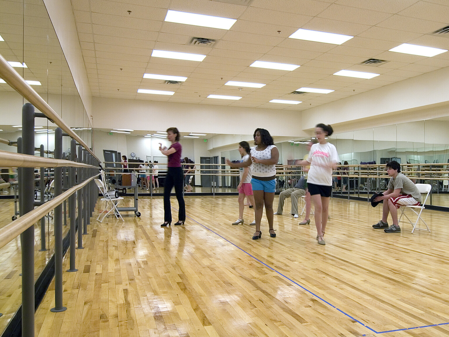 Dance room with mirrors on all walls, natural wood floors, filled with students learning dance