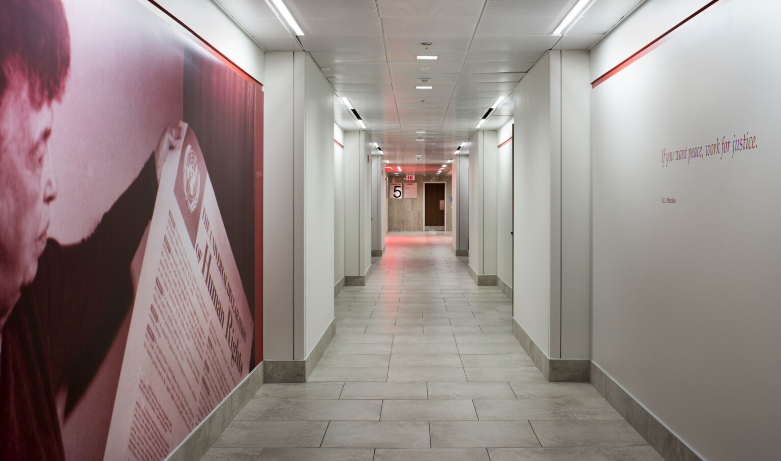 White hallway with red accent stripes. Front left section of hall has photo of a man with a piece of paper, opposite is quote