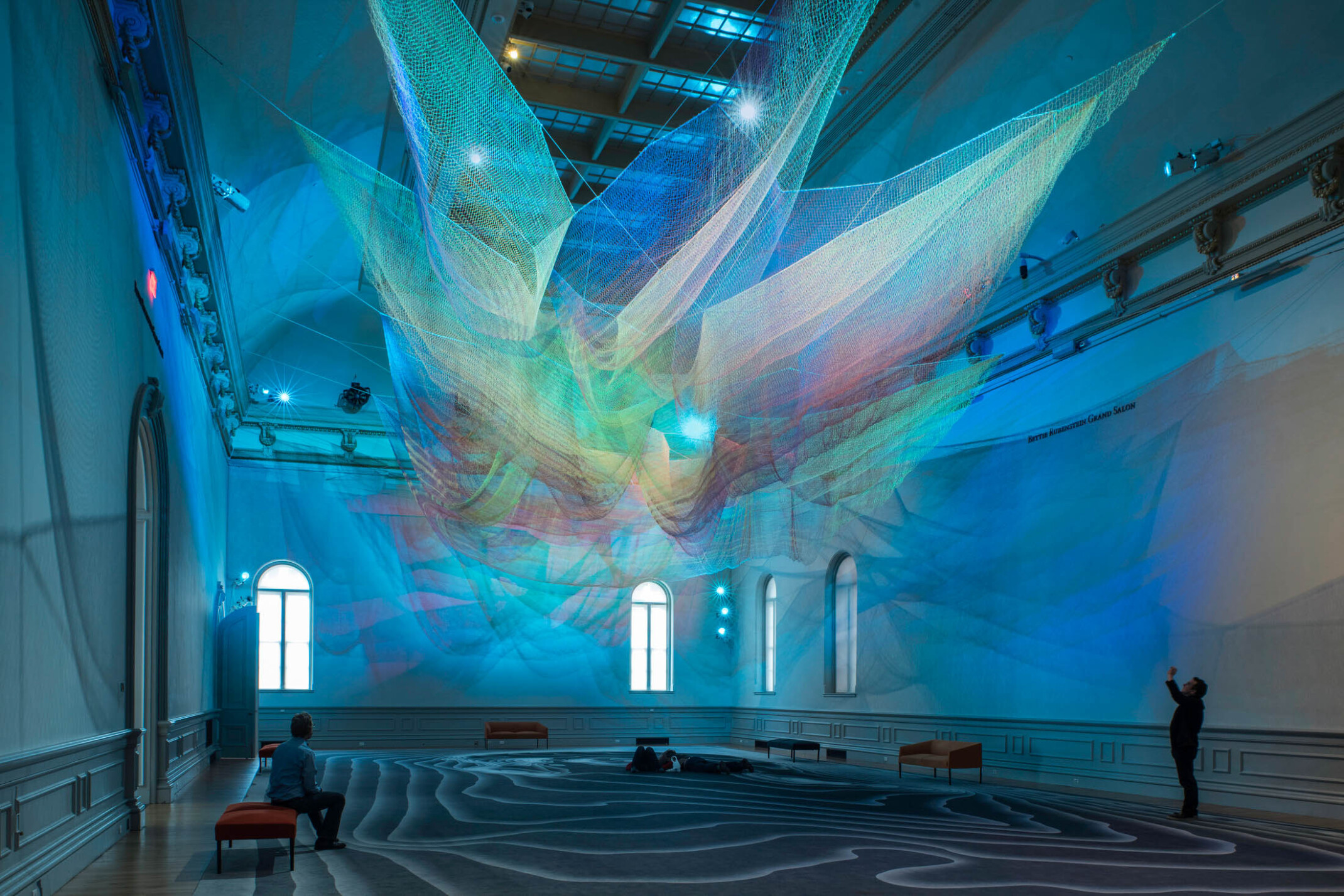 Colorful abstract art installation with green and blue light on layers of sheer netting with shadows and light on white wall
