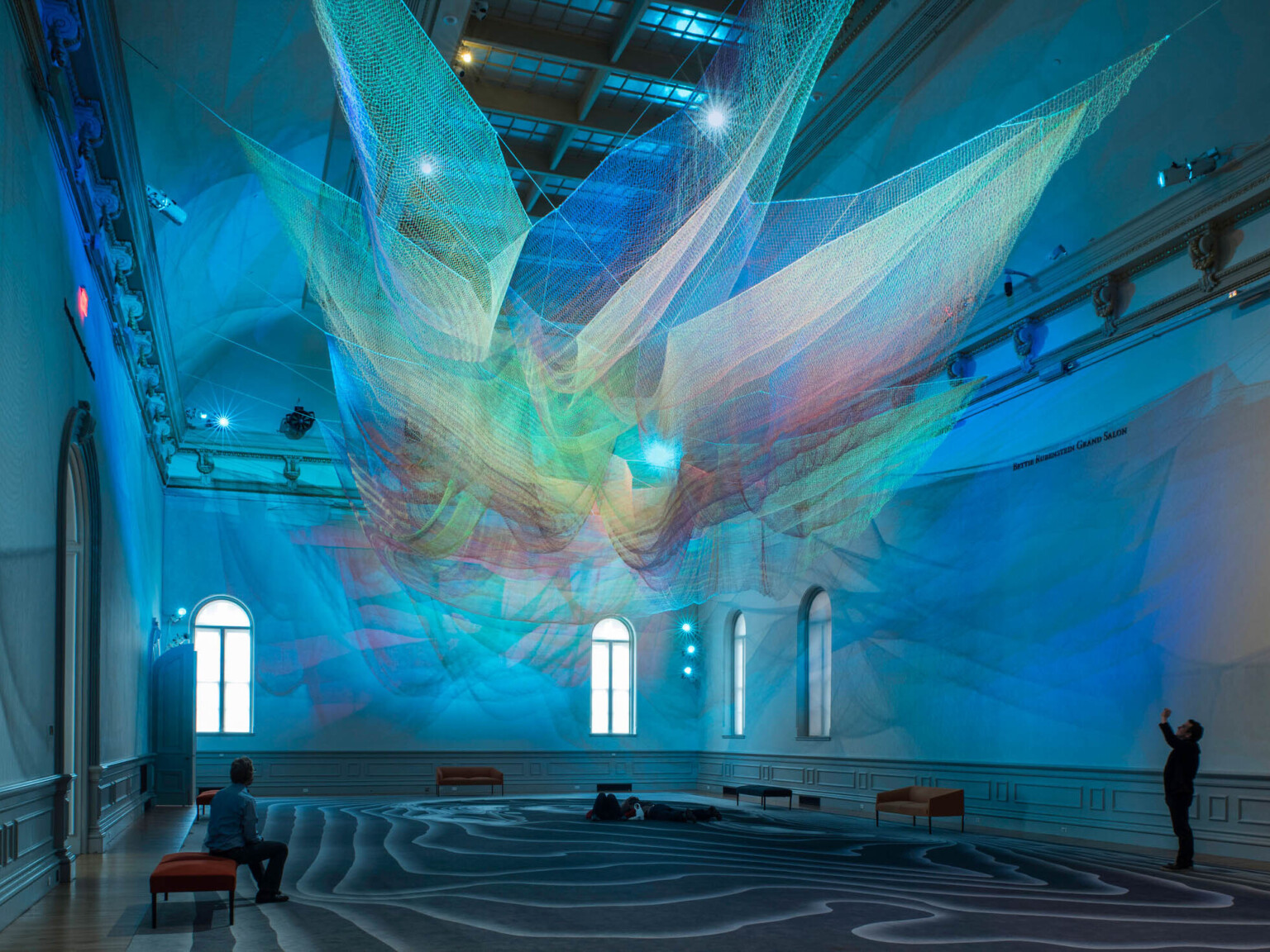 Colorful abstract art installation with green and blue light on layers of sheer netting with shadows and light on white wall