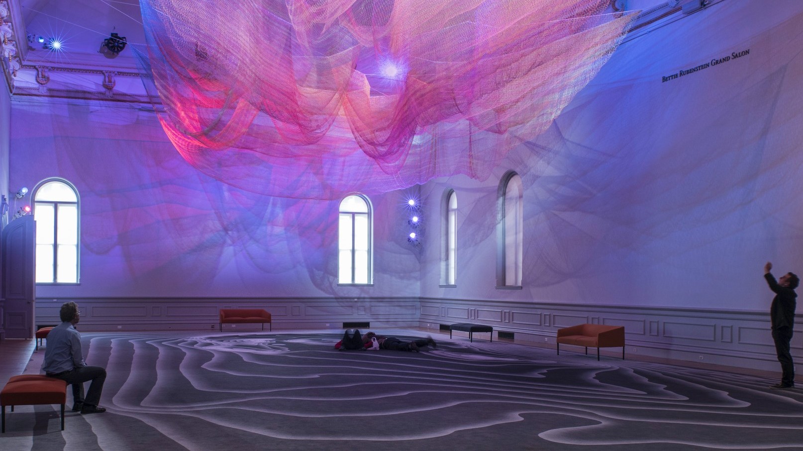 Colorful abstract art installation with pink and purple light on layers of sheer netting with shadows and light on white wall